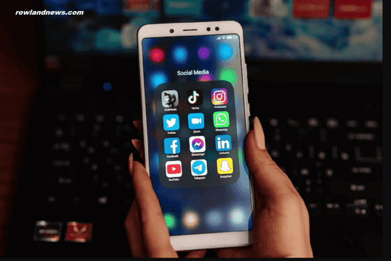 A person holds a smartphone displaying various social media app icons, including TikTok and Instagram, suggesting the ability to install Android Apps on iPhone, with a blurred laptop in the background.