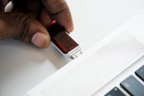 A hand is inserting a red USB flash drive into the USB port of a white laptop. The laptop keyboard and speaker grille are partially visible.